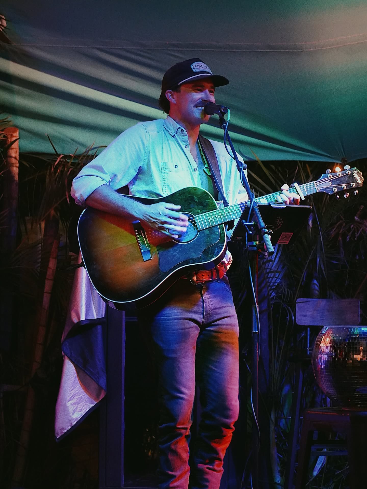 Pierce August singing into the mic while playing guitar under a tent stage